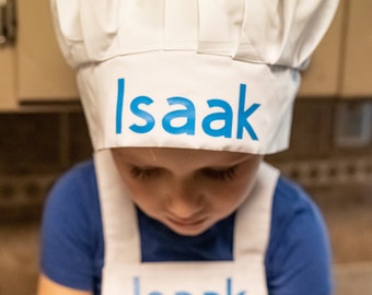 May include: A young child wearing a white chef hat and apron with the name "Isaak" printed in blue on both. The child is looking down.