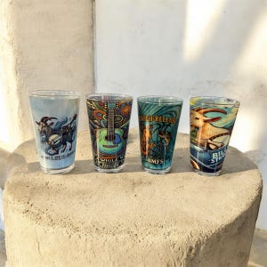 May include: Four clear pint glasses with colorful designs. One glass features a goat playing a banjo, another a guitar, and a third with fish and the words "CATCH-RELEASE". The last glass has a goat and the words "BILL STR".