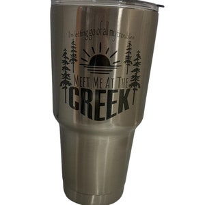 Puede incluir: Un vaso de acero inoxidable con un gráfico negro que dice "Me estoy deshaciendo de todos mis problemas... Encuéntrame en el arroyo". El gráfico también incluye una puesta de sol y pinos.