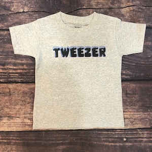 May include: A light gray t-shirt with the word "TWEEZER" in a black and blue font. The shirt is made of a soft material and is suitable for children. The shirt is displayed on a wooden surface.
