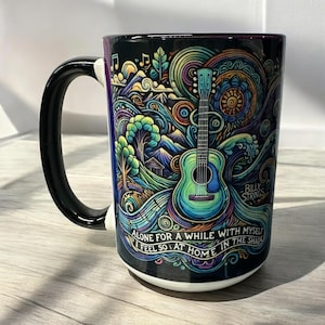 Puede incluir: Una taza de café negra y blanca con un diseño colorido que presenta una guitarra, montañas y árboles. El texto "Alone for a while with myself I feel so at home in the shade" está impreso en la taza.