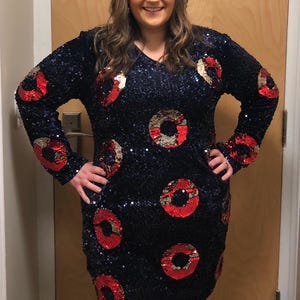 May include: A long-sleeved, navy blue sequined dress with a V-neck. The dress features large, circular patterns in red and gold sequins. The dress is knee-length and appears to be made of a shimmering fabric, suitable for a special occasion.