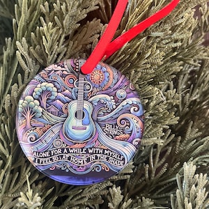 May include: Round Christmas ornament with a colorful, abstract design featuring a guitar, waves, and a house. The ornament has a red ribbon and the text "BILLY STRINGS ALONE FOR A WHILE WITH MYSELF I FEEL SO AT HOME IN THE SHADE".