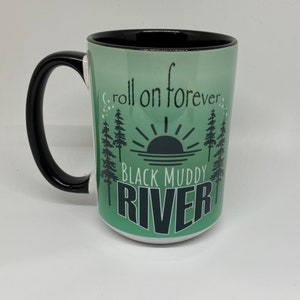 May include: A green and black ceramic mug with a sunrise and trees design. The text "Roll on forever Black Muddy River" is printed on the mug.