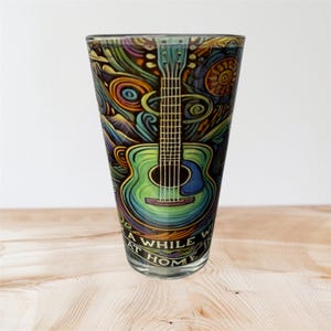 Puede incluir: Un vaso alto y transparente con un diseño psicodélico colorido que presenta una guitarra verde. La guitarra es el foco central, rodeada de patrones arremolinados en tonos de azul, verde, amarillo y morado. El texto en la parte inferior dice "A WHILE AT HOME".