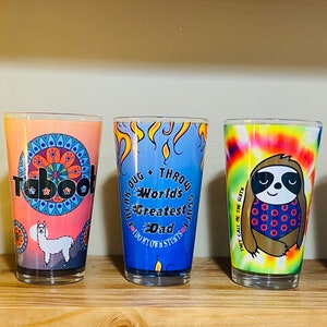 Puede incluir: Cinco tazas de vidrio de colores diferentes con varios diseños. Las tazas presentan una taza amarilla con un diseño de ciervo y el texto "OUT OF CONTROL", una taza rosa con un diseño de llama y el texto "Taboot", una taza azul con un diseño de llama y el texto "JUG + THROW World's Greatest Dad BUY MY OWN STUFF", una taza tie-dye con un diseño de perezoso y el texto "THEY CALL ME THE DADDY", y una taza azul con un diseño de figuritas de palo y el texto "MEATSTICK".