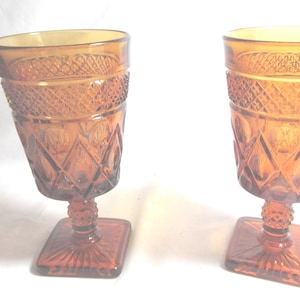 May include: Two amber-colored vintage goblets. The glasses have a diamond pattern around the top and diamond and oval patterns on the body. The goblets sit on a square base with a textured stem.