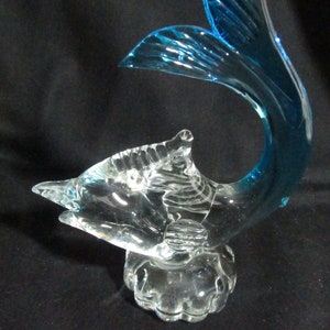 Puede incluir: Una escultura de vidrio transparente de un pez con una aleta caudal azul. El pez est&aacute; de pie sobre una base de vidrio transparente.
