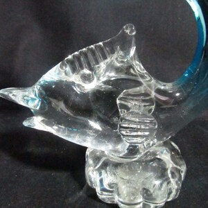 Puede incluir: Una escultura de cristal transparente de un delf&iacute;n con una aleta caudal azul. El delf&iacute;n est&aacute; enroscado en un c&iacute;rculo y tiene un cuerpo texturizado. La escultura est&aacute; sobre una base de cristal transparente.