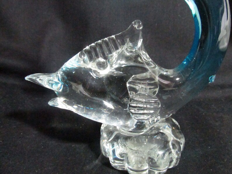 Puede incluir: Una escultura de cristal transparente de un delf&iacute;n con una aleta caudal azul. El delf&iacute;n est&aacute; enroscado en un c&iacute;rculo y tiene un cuerpo texturizado. La escultura est&aacute; sobre una base de cristal transparente.