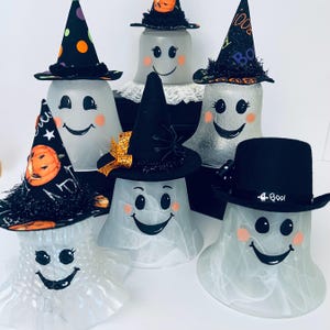 May include: A collection of five translucent ghost figurines, each wearing a unique miniature hat. The ghosts feature painted black eyes, wide smiling mouths, and orange circular cheeks. The hats include patterns of pumpkins, spiders, and the word Boo.