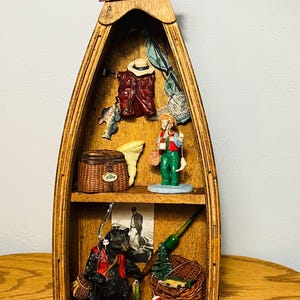 May include: A wooden boat-shaped wall shelf with two tiers, featuring a sign at the top that reads BORN TO FISH. The display contains miniature fishing-themed figurines, including a person in green overalls, a black bear, wicker baskets, and nets.