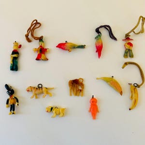 May include: A collection of 12 vintage plastic charm pendants. The charms include a clown, a bird, a parrot, a boy, a tiger, a giraffe, a banana, a corn, a dog, a rabbit, a doll, and a sailor. The charms are colorful and have a retro look.