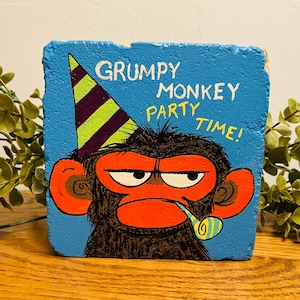 May include: A square blue stone painted with a black monkey wearing a purple and lime green striped party hat. The monkey has a bright orange face, a party blower in its mouth, and the text GRUMPY MONKEY PARTY TIME! written in white and yellow.