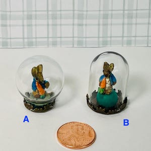 May include: Two miniature glass cloches, each containing a figurine of a rabbit wearing a blue jacket and holding a carrot. One is in a round glass dome, the other in a bell-shaped dome. A penny is in the foreground.