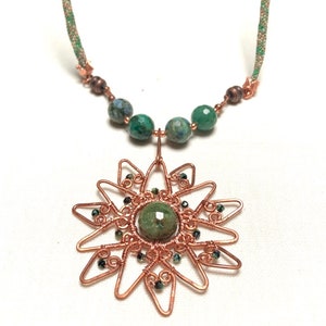 May include: A handcrafted copper wire necklace featuring a sunburst pendant with a central green stone. The necklace includes green and blue beaded accents and a braided cord. The pendant is approximately 2 inches in diameter.