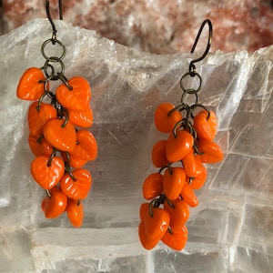 Puede incluir: Un par de pendientes en forma de corazón naranja. Cada pendiente presenta un grupo de cuentas brillantes en forma de corazón suspendidas de un gancho de color bronce. Los pendientes están colocados sobre un fondo texturizado y claro.