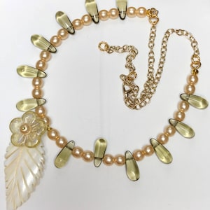 May include: A delicate necklace featuring peach-colored faux pearls, teardrop-shaped amber-toned beads, and a carved leaf pendant. The necklace includes a flower-shaped accent and a gold-toned chain with a lobster clasp.