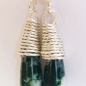 May include: A pair of handcrafted dangle earrings. Each earring features a teardrop-shaped green and white stone wrapped with silver-colored wire. The wire is tightly coiled around the stone and forms a loop at the top for the earring hook.