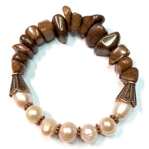 Könnte beinhalten: Ein Stretch-Armband mit braunen, polierten Steinperlen und großen, cremefarbenen Süßwasserperlen. Das Armband hat kupferfarbene Metallakzente. Die Perlen haben einen zarten, rosafarbenen Farbton.