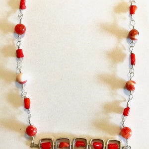 May include: A handcrafted necklace with red coral beads and silver-tone accents. The necklace features a toggle clasp and is strung with a mix of round and cylindrical coral beads. The design includes square silver settings with red coral pieces and dangling coral.