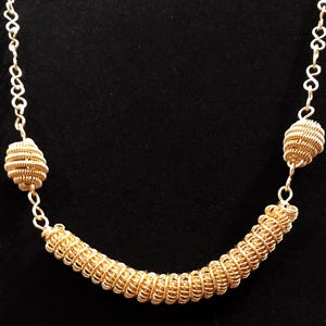 Può includere: Collana color oro dal design unico. La collana presenta un elemento centrale curvo in filo avvolto e due perline decorative a forma di spirale. La catena è composta da piccoli cerchi collegati. Il gioiello è esposto su sfondo nero.