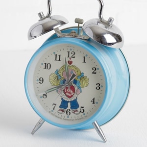 May include: A blue alarm clock with silver bells and a white face. The clock face features a cartoon girl with blonde pigtails holding a heart that says "I love you". The clock face also has black numbers and hands.