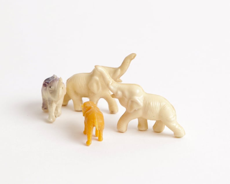 May include: A set of four toy elephants, three are white and one is purple. The elephants are standing on a white surface. There is also a small orange toy elephant in the center of the image.