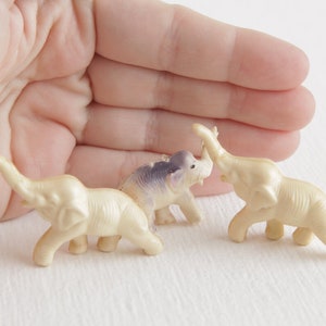 May include: Three small plastic toy elephants, one white, one purple, and one white with a raised trunk. The elephants are arranged in a semi-circle on a white surface.