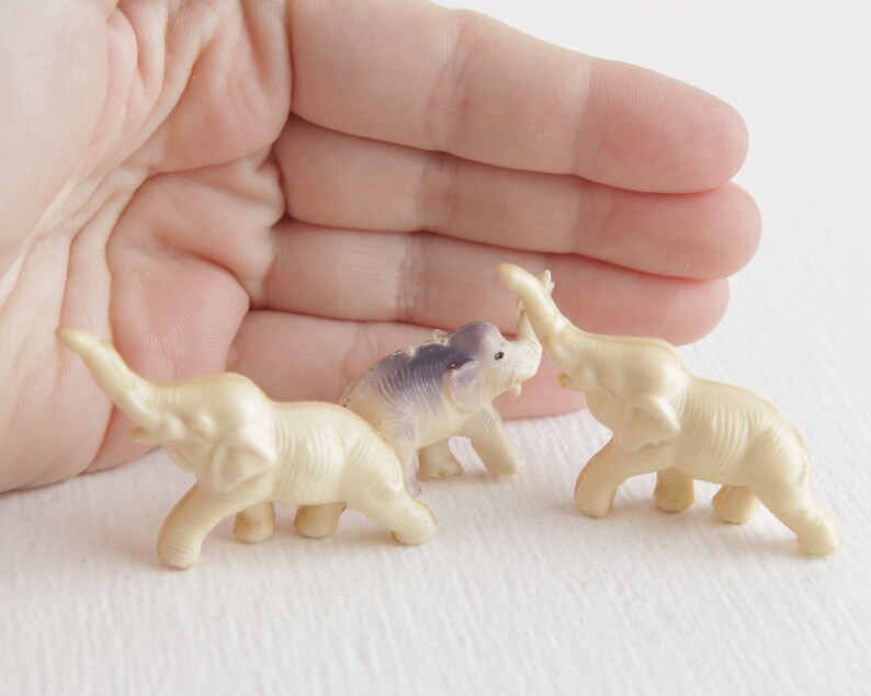 May include: Three small plastic toy elephants, one white, one purple, and one white with a raised trunk. The elephants are arranged in a semi-circle on a white surface.