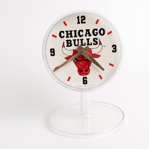 May include: A white desk clock with a red and black Chicago Bulls logo. The clock has black numbers and red tick marks. The clock is on a clear plastic stand.