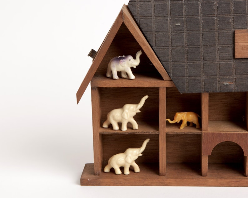 May include: A wooden house-shaped shelf with four compartments, each containing a small elephant figurine. The elephants are white, purple, and orange.
