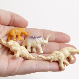 May include: Four small plastic toy elephants in a hand. One elephant is orange, and the other three are cream colored. The elephants are all facing different directions.