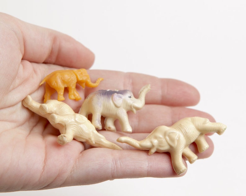 May include: Four small plastic toy elephants in a hand. One elephant is orange, and the other three are cream colored. The elephants are all facing different directions.