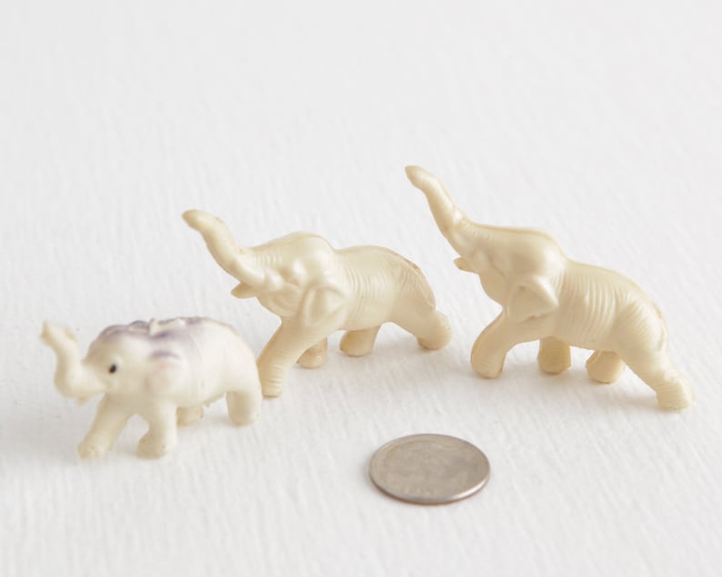 May include: Three small, white, plastic toy elephants. The elephants are in a line, with the one in the back facing forward, the middle elephant facing left, and the front elephant facing right.
