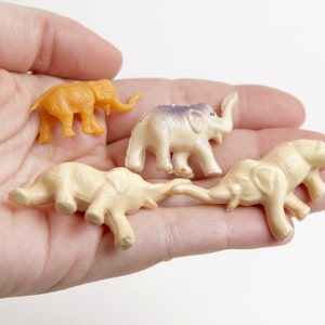 May include: Four small plastic toy elephants in a hand. One elephant is orange, and the other three are cream colored.