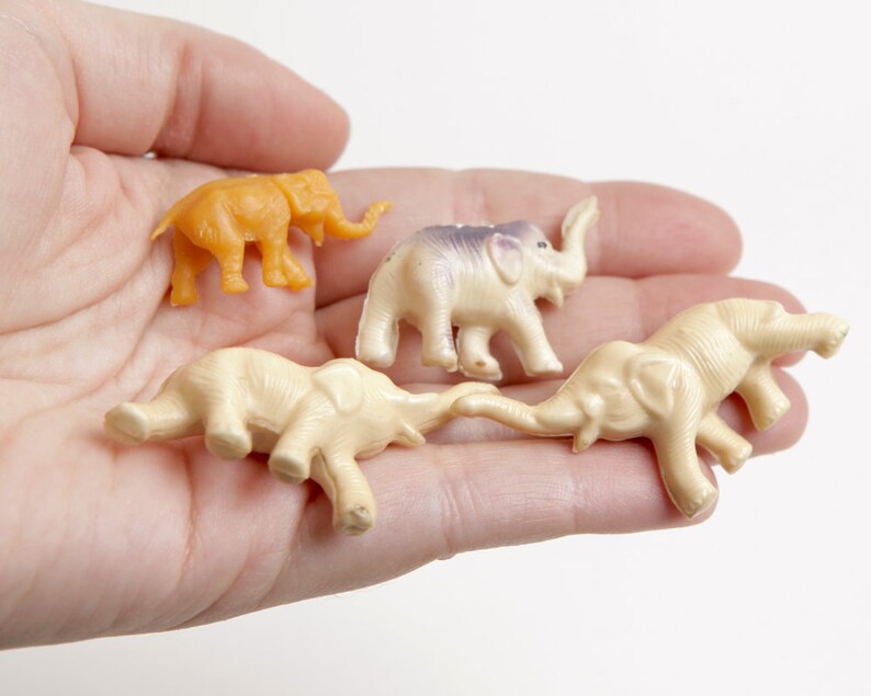 May include: Four small plastic toy elephants in a hand. One elephant is orange, and the other three are cream colored.