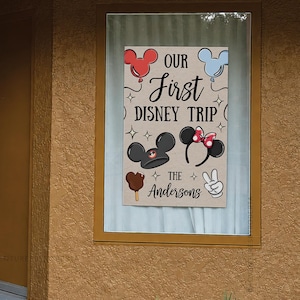 May include: A rectangular sign with the text "OUR First DISNEY TRIP" and "The Andersons" in a whimsical font. The sign features Mickey Mouse ears, a Mickey ice cream bar, and a peace sign.