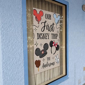 May include: A framed sign with the text "OUR First DISNEY TRIP" and "THE Andersons". The sign features Mickey Mouse and Minnie Mouse ears, a Mickey ice cream bar, and a peace sign. The sign is displayed in a window with a blue frame.