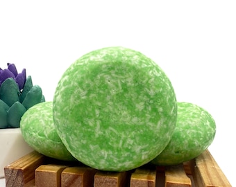 May include: Three round, green shampoo bars with a textured surface, resting on a small wooden soap dish. The bars have a light green color with white flecks. A decorative succulent is visible in the background.