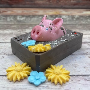 May include: A pink pig with black eyes and a white nose is lying on a dark gray soap bar. The soap bar is decorated with white sprinkles and blue and yellow flowers.