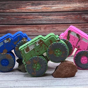 May include: Three colorful toy monster trucks in blue, green, and pink, with large black wheels and brown accents. The trucks are arranged on a light wood surface, with a dark wood background.