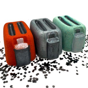 May include: Three colorful, novelty toaster-shaped objects. One is orange, one is silver, and one is teal. Each has a black slot and a black, rectangular base. The objects are on a white surface with black confetti.