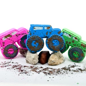 May include: Three truck-shaped bath bombs in bright pink, blue, and green. Each vehicle features large, dark-colored wheels and is decorated with brown, mud-like splatters across the body and tires. The bath bombs are arranged on small rocks.