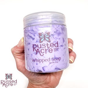 A jarsof purple lilac whipped soap handmade by Rusted Acre Soap Co. in a hand.