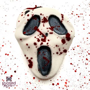 May include: A white bath bomb shaped like a Scream mask, with dark gray eye and mouth openings. Red splatters resembling blood are on the mask and surrounding white surface. The product is from Rusted Acre Soap Co.