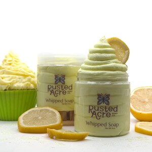 May include: Two clear plastic jars of pale yellow whipped soap labeled Rusted Acre Soap Co. with a butterfly logo. One jar features a tall swirl of soap topped with a lemon slice. A green cupcake liner with yellow soap and lemon slices are nearby.