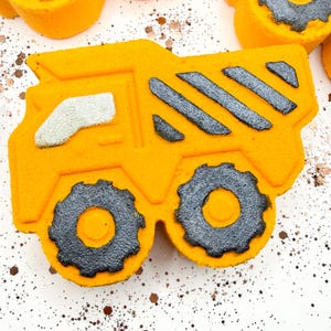 May include: A bright orange dump truck shaped bath bomb featuring a silver glittery window, two large black glittery textured wheels, and diagonal silver glittery stripes on the truck bed. The surface is matte orange with sparkling accents.