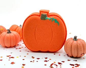 May include: A large, flat, orange pumpkin-shaped bath bomb with a green stem detail, surrounded by several smaller, textured, orange pumpkin-shaped bath bombs and scattered red and orange leaf-shaped glitter pieces on a white surface.