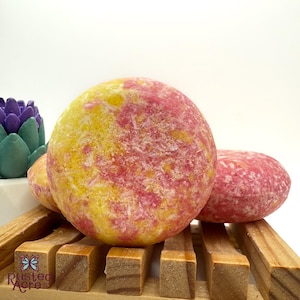 May include: Two pink and yellow solid shampoo bars on a wooden rack. The bars have a speckled appearance.